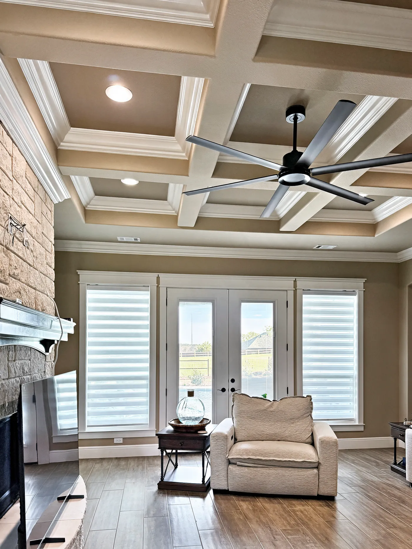 Zebra shades on windows beside French doors in a living room with a coffered ceiling and stone fireplace